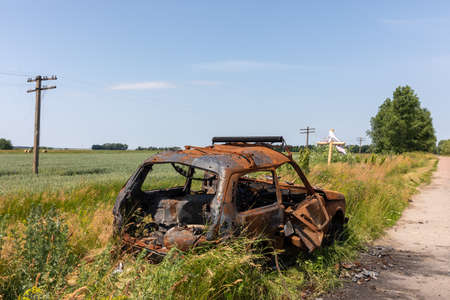 CHERNIHIV REG, UKRAINE - Jun. 19, 2022: A burned-out car on the side of the road against the background of a wheat field and a grave cross. Landscapes of Ukraine after the invasion of Russian fascistsのeditorial素材