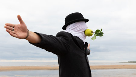 A man with a face covered by a cloth, wearing a hat and a coat, is holding an apple in his mouth without using his hands on the seashore.の写真素材