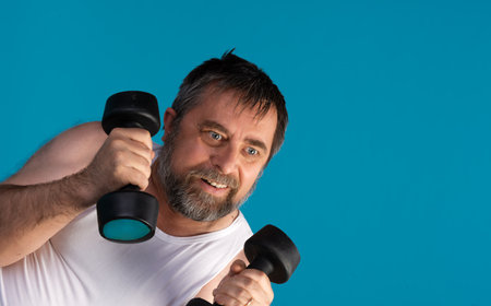 A middle-aged man with a beard lifts dumbbells in a vibrant blue studio, showcasing his determination and strength during an intense workout session aimed at improving fitness.の写真素材