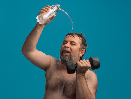 A dedicated elderly man focuses on his workout as he curls dumbbells with one hand while splashing water on his head with the other hand, enjoying a refreshing moment.の写真素材