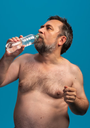 A middle-aged man drinks water from a bottle while taking a break from sports. He appears relaxed and focused on rehydrating after physical exertion, enjoying the moment.の写真素材
