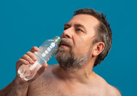 A middle-aged man drinks water from a bottle while taking a break from sports. He appears relaxed and focused on rehydrating after physical exertion, enjoying the moment.の写真素材