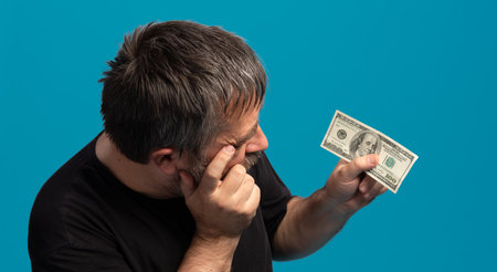 A man is closely inspecting a one hundred dollar bill, demonstrating curiosity and focus. His expression hints at interest while he holds the bill against a vivid blue backdrop.の写真素材
