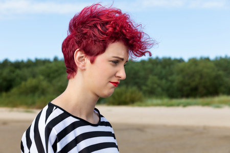 A young woman with vibrant red, curly hair strolls along a sandy area. She wears a black and white...