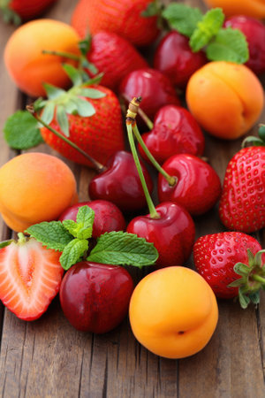 An assortment of colorful summer fruits including cherries, strawberries, apricots, and mint leaves is neatly spread on a rustic wooden surface. The fruits display vivid colors and textures.の写真素材