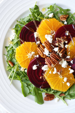 A vibrant salad features fresh arugula topped with orange slices, roasted beets, crumbled cheese, and toasted pecans. A drizzle of dressing completes the dish presented on a white plate.の写真素材