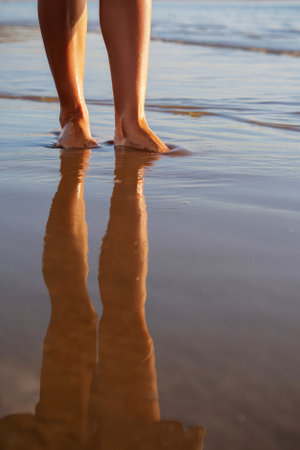 Feet stand in shallow water at a peaceful beach during twilight. Gentle waves reflect on the wet sand, creating a tranquil atmosphere where nature and serenity unite.の写真素材