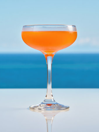 A cocktail filled with a vibrant orange liquid sits elegantly in a tall glass on a reflective surface, with a beautiful ocean backdrop and clear blue sky creating a relaxing atmosphere.の写真素材