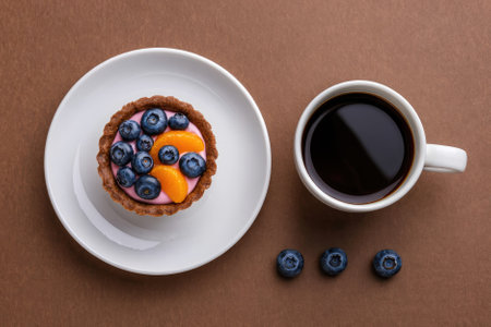 A delightful dessert consisting of a colorful tart topped with fresh blueberries and orange slices, paired with a steaming cup of coffee on a brown table.の写真素材