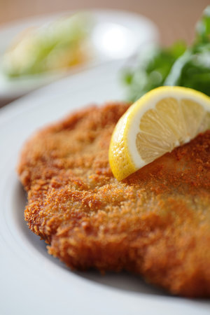 A crispy breaded cutlet is plated with a slice of lemon perched on top, accompanied by a side of...