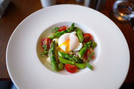 A vibrant dish featuring a perfectly poached egg atop a mix of cherry tomatoes, asparagus, and greens, showcasing fresh ingredients artfully arranged in a bowl.の写真素材