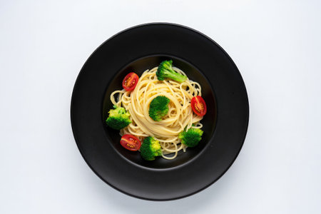 A top-down shot showcases a black plate filled with pasta, broccoli, and cherry tomatoes on a white background.の写真素材