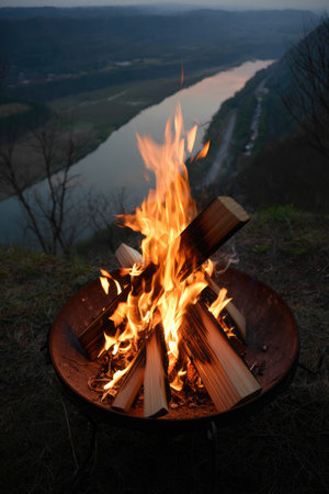 A warm fire burns in a metal pit overlooking a vast river valley as dusk settles over the landscape.