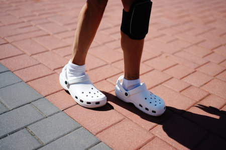 A person wearing white clogs and a black device on the leg stands on a brick paved surface.