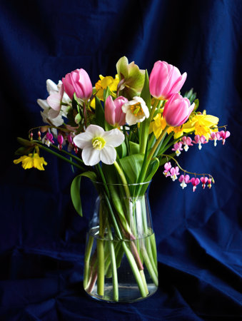 A vibrant bouquet of spring flowers in a glass vase, showcasing a variety of colorful blooms against a dark blue backdrop.の写真素材
