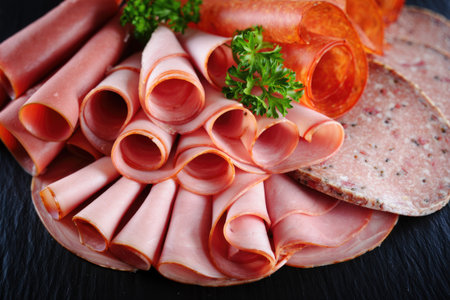 A close up of various sliced deli meats arranged on a dark surface, including ham, salami, and pepperoni.の写真素材