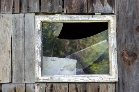 A close up view of a broken window in an old wooden building. The frame is weathered, and the glass...