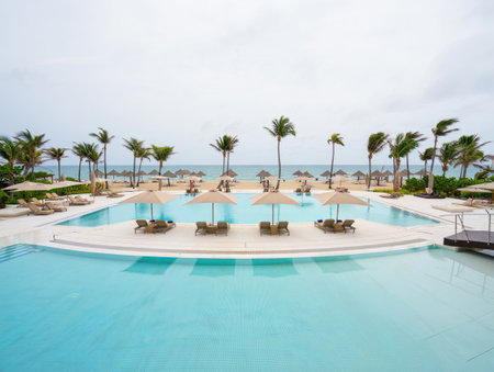 A luxurious resort scene with a pool, beach, and palm trees on a cloudy day, evoking relaxation and...