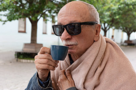 An elderly man wearing sunglasses drinks from a blue mug while wrapped in a blanket, enjoying a...