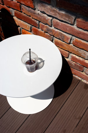 A refreshing iced coffee sits on a white table with a rustic brick wall background. The composition highlights the contrast and detail.の写真素材