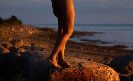 A person's legs stand on a sunlit rock by the coast with the sea in the background. The warm light bathes the scene, creating a serene atmosphere.の写真素材