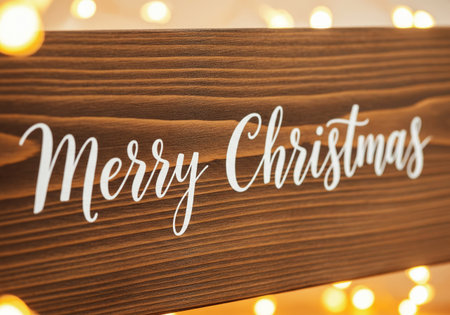 A wooden sign with the words Merry Christmas in elegant white script is displayed with warm, glowing lights in the backgroundの写真素材