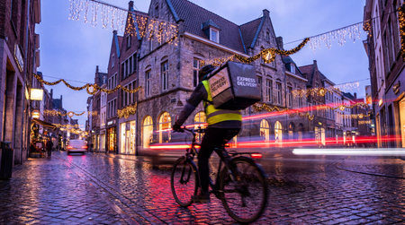 Blurred delivery cyclist speeds along a festively lit cobblestone street, carrying an express...
