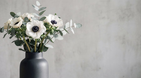 A bouquet of white anemone flowers with dark centers and eucalyptus leaves is arranged in a matte...