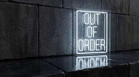 A white neon sign displaying the words OUT OF ORDER sits inside a clear glass case. The sign leans against a dark, textured stone wall. Cool light reflects on the wet floor below.の写真素材