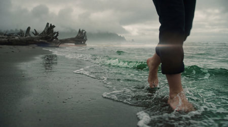 Lower legs and bare feet of an individual walking along the shoreline where small green-tinged waves wash over dark, wet sand, with large driftwood logs visible in the background under an overcast.の写真素材