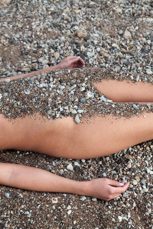The lower torso and legs of a person lie on a coarse, multi-colored pebble beach. Sand and small stones cover the midsection, contrasting with exposed skin on the arms and thighs.の写真素材
