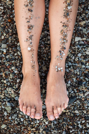 Two bare legs and feet rest vertically on a surface covered in small, multi-colored pebbles. Sand and small stones adhere to the front of the legs in vertical lines.の写真素材