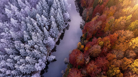 An aerial view divides a river between a forest covered in white frost or snow on the left and a...