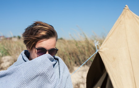Young woman near the tent on the beach covers her face from the wind
