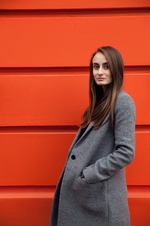 Close up portrairt of young beautiful woman with brunette hair wearing grey coat on the orange wall.の写真素材