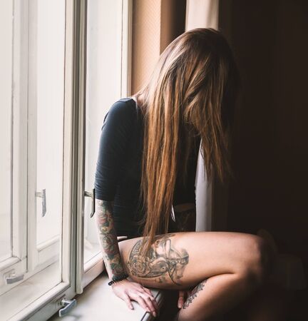 Young tattooed woman with long hair sitting on the windowsill, wearing black bodysuitの写真素材