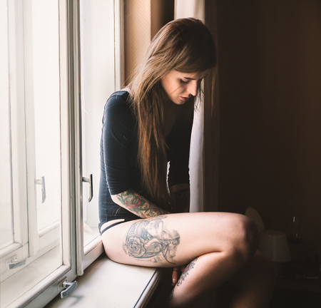 Young tattooed woman with long hair sitting on the windowsill, wearing black bodysuitの写真素材