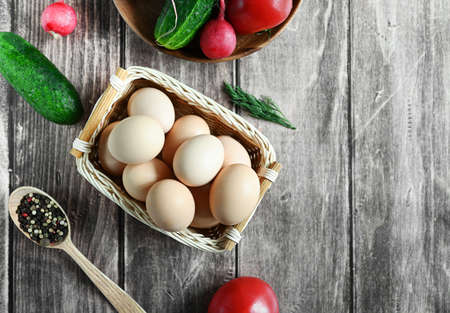 Fresh homemade beige eggs in a basket and vegetables on a wooden table, top view, copy space.
