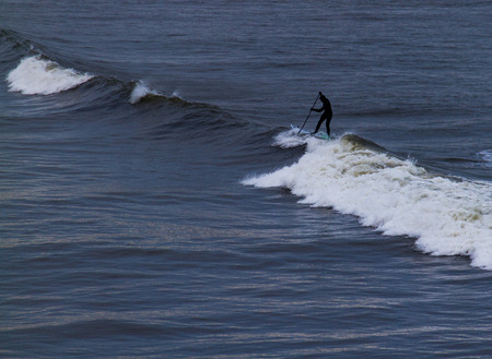 Surfer holding oar in wetsuit negotiating waves  on Tynemouth beach in UK on a winter morning