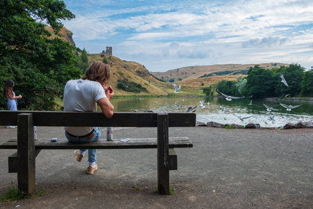Edinburgh, United Kingdom - July 27, 2018: Young man sits and smokes on a bench by St Margaretâs Loch with ruins of St Anthonyâs chapel in the background.のeditorial素材
