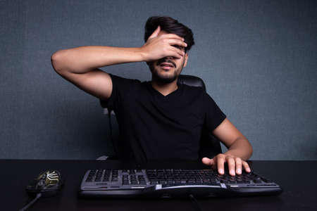 the young man covered his face with his hand. Sits in front of the monitor. On a gray backgroundの写真素材