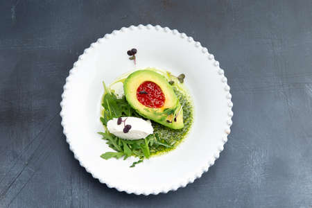 Snack. Avocado with red caviar and arugula in a plate. On a gray background. Selective focus.