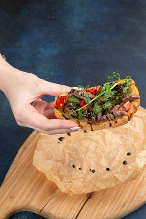 Hand holds a bruschetta with meat, tomatoes and herbs. Against a dark background. Selective focus.の写真素材