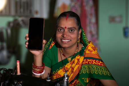 Indian rural woman using sewing machine and SHOWING HER MOBILE PHONEの写真素材