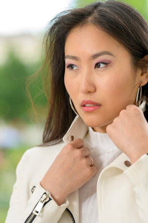 A young pretty Mongolian woman with a white leather jacket in an urban park.