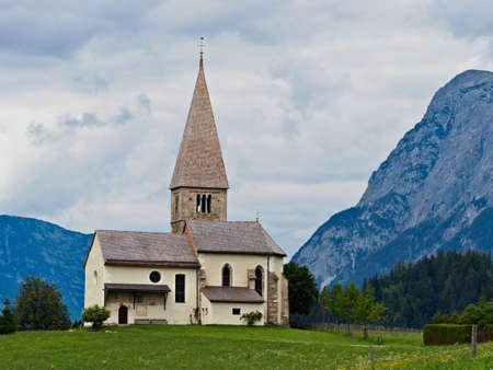 A summer meadow with a blue sky and a few white clouds with a large mountain range in the background. A small chapel with a gnarled fruit tree can be seen in the foreground.の写真素材