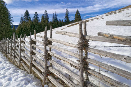 An old wooden fence that is held together with willow rods and separates two snowy pastures from each other. In addition, the fence is reinforced with a barbed wire.の写真素材