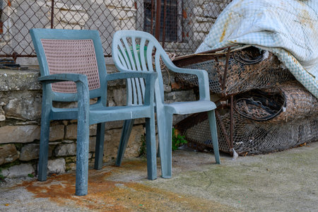 Two weathered plastic chairs sit next to a pile of rusty metal mesh and a discarded blanket. Rust stains the ground.の写真素材