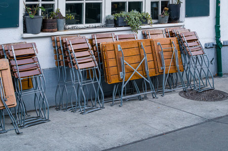 Folded wooden tables and chairs are stacked against a building with potted plants on the window...