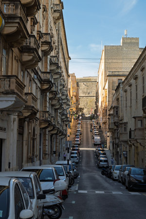 A steep street in Valletta, Malta, is lined with parked cars and pedestrians. The architecture is...